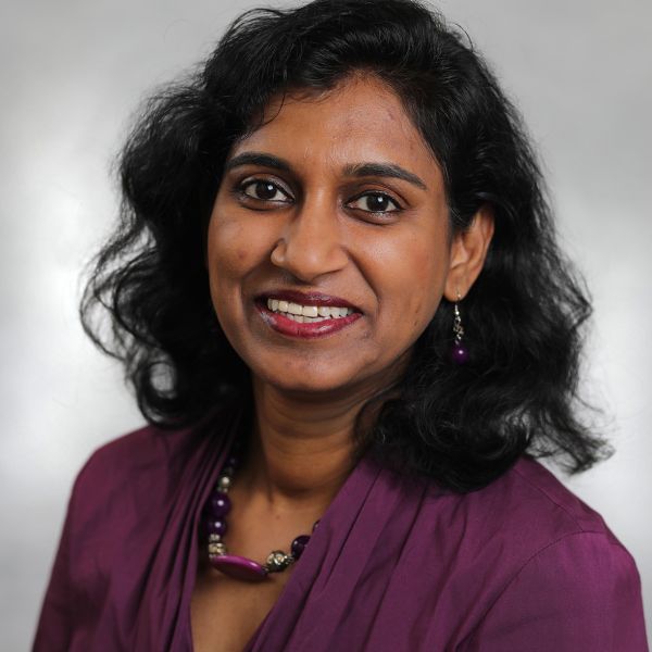 A smiling Preethi Mony with short wavy black hair and wearing a purple blouse
