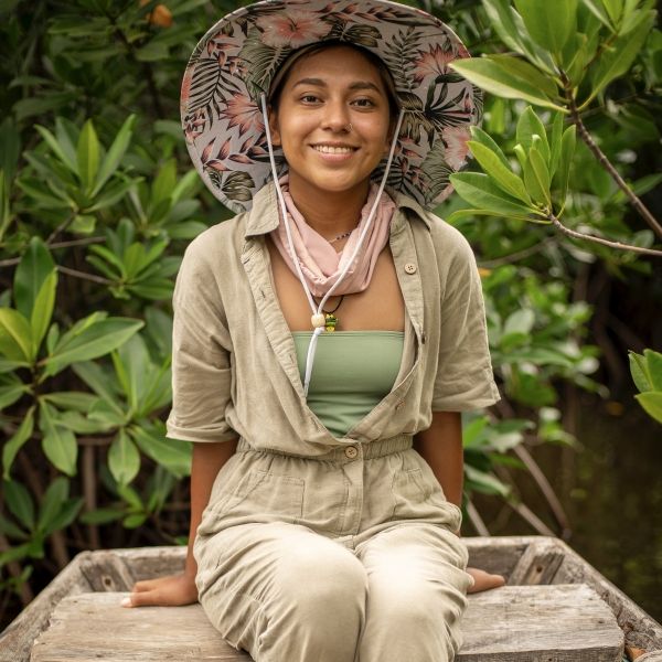 EE 30 Under 30 Awardee Jabel Gomez sits on a wooden bench while wearing a sun hat and sandals, green plants behind her