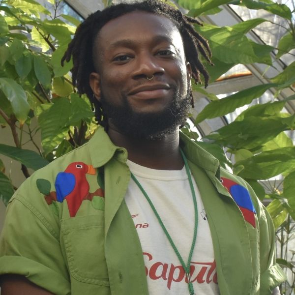 EE 30 Under 30 Awardee Jibreel Cooper wearing an open green button-up with parrots on the shoulders, standing in front a green leafy plant