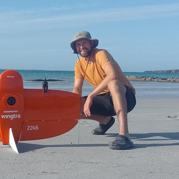 EE 30 Under 30 Awardee Joe Boyle crouches on a beach with a blue sky and the ocean behind him