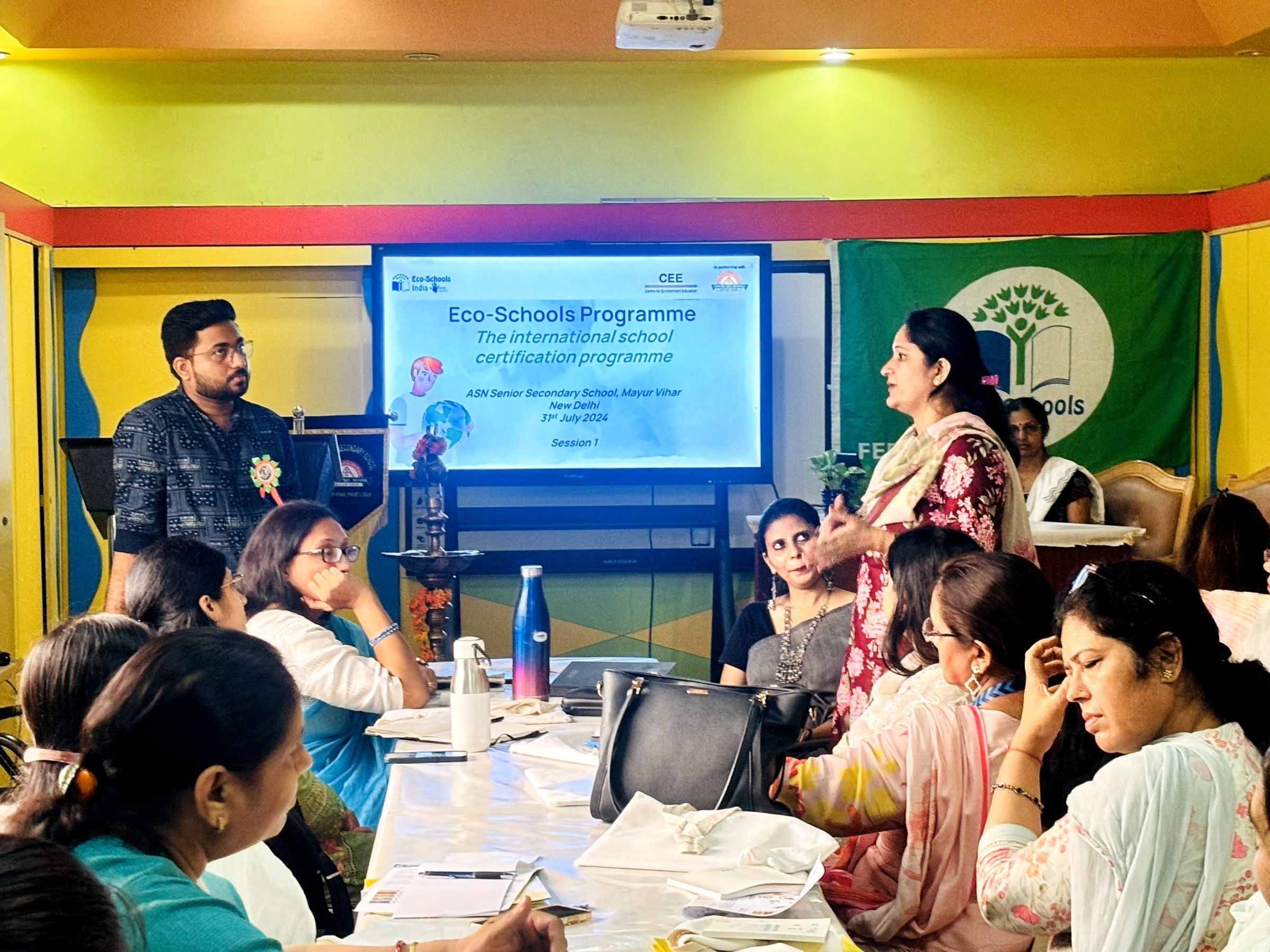 Two speakers in front of a group of seated people with a projector that reads, "Eco Schools Programme."