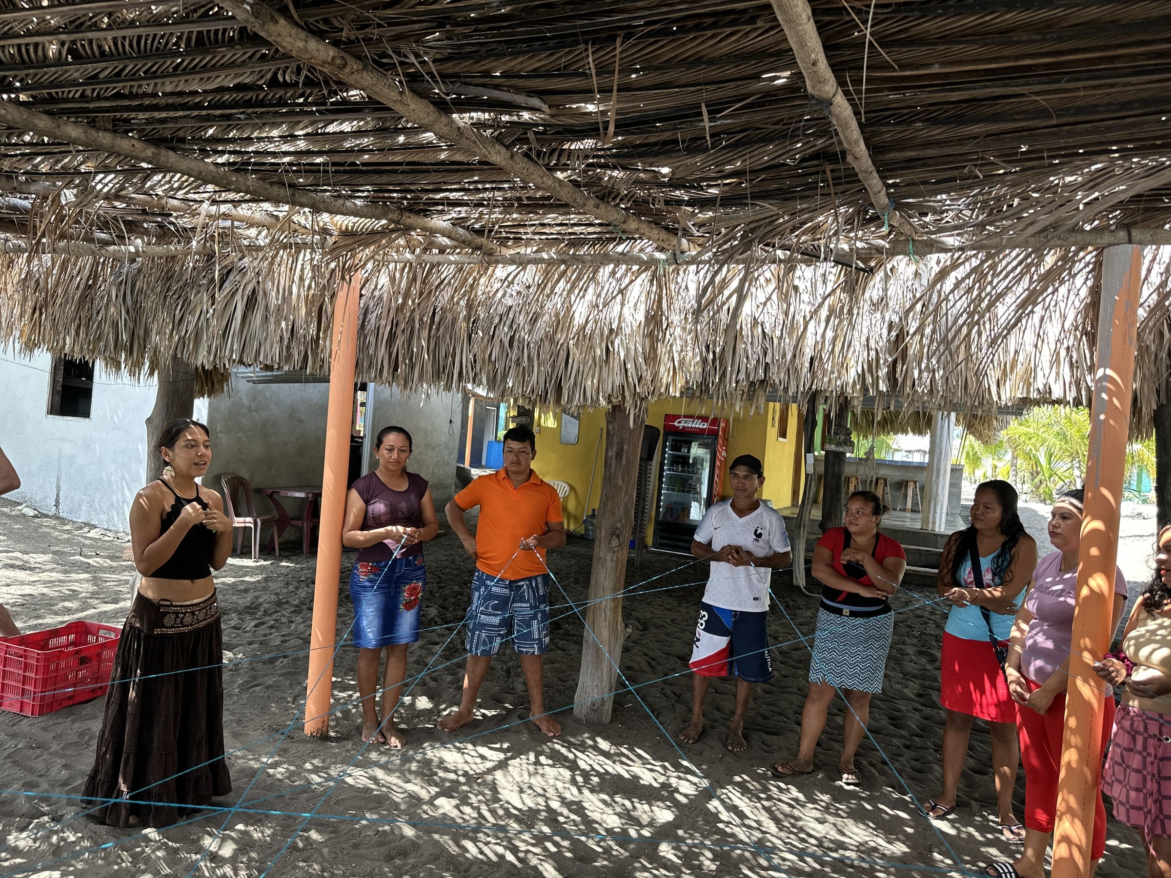 A group of people under a dried leafy canopy outdoors hold blue string between them