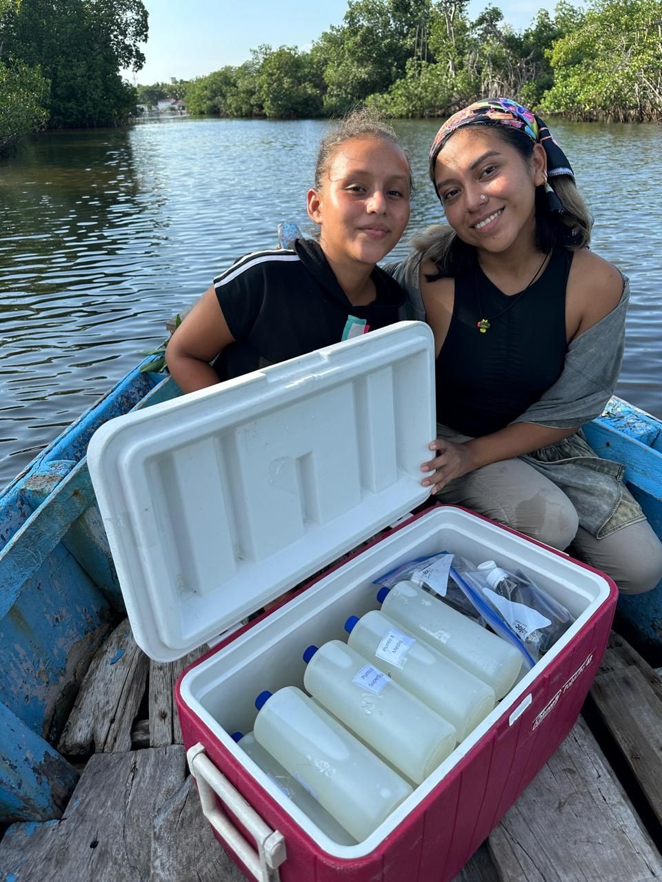 A child and person sit in a boat with water and green foliage behind them; in front of them is a cooler full of bottles