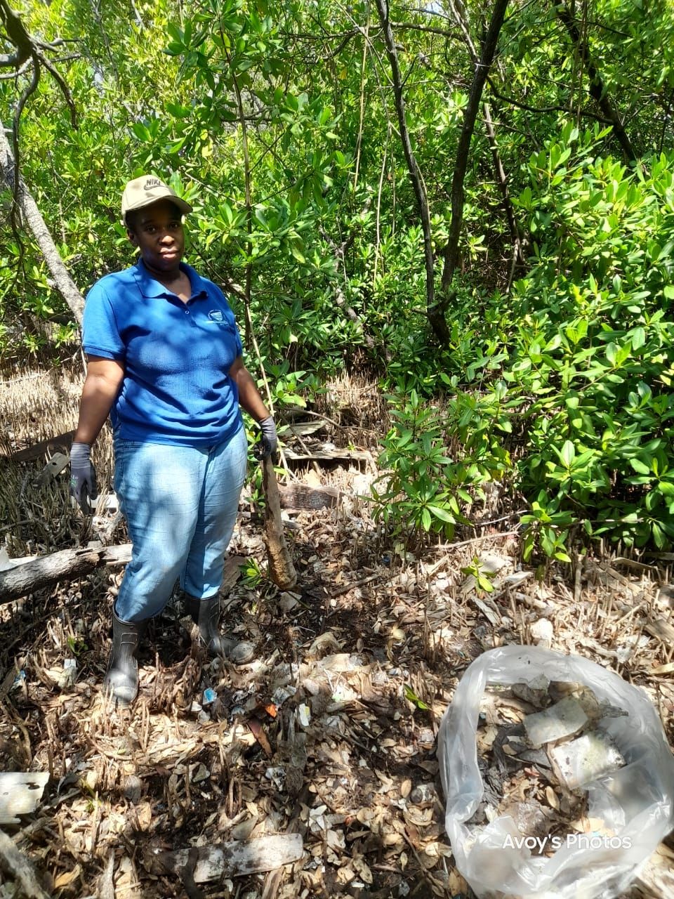CEE-Change Fellow Nicole in a blue shirt and cap within a heavily wooded area, holding a stick in one gloved hand.
