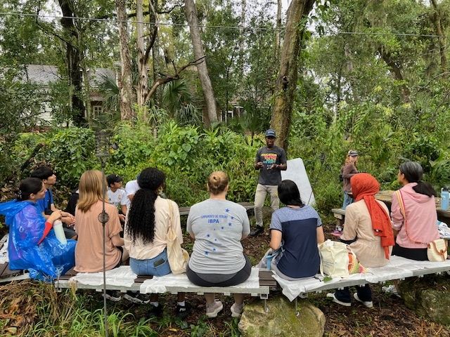 CEE-Change 2025 Fellow Jeftey presents to a group of people sitting on benches in a heavily forested area.