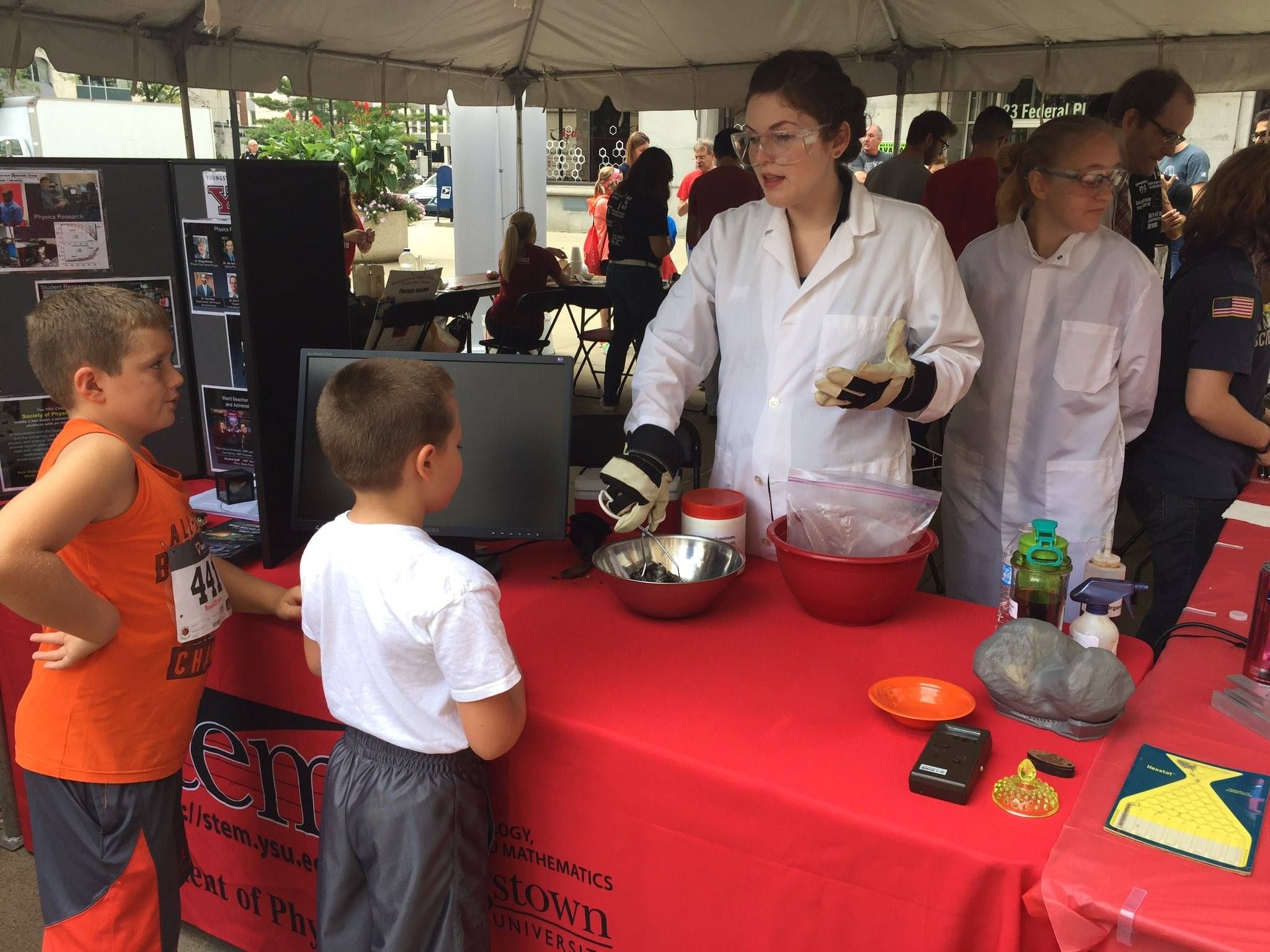CEE-Change 2025 Fellow Tiffany stands behind a booth wearing a white labcoat, demonstrating science to two children.