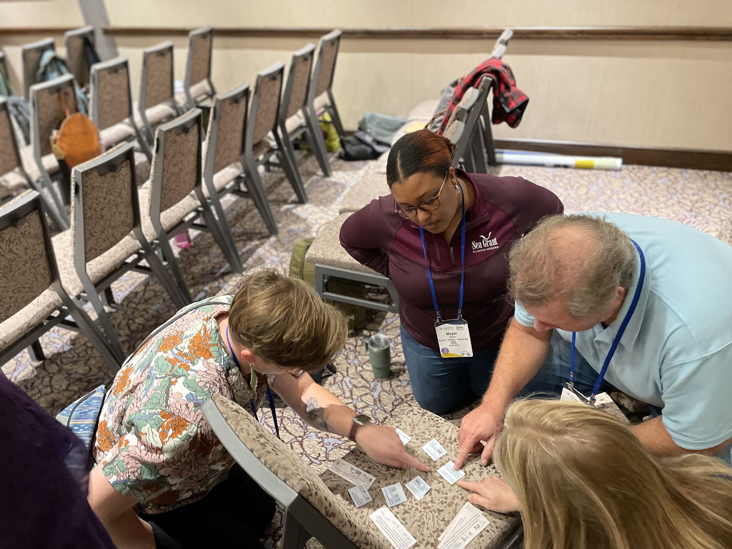 Four conference attendees kneel around a chair in a meeting room, collaborating on a hands-on activity by arranging and discussing cards spread across the seat.