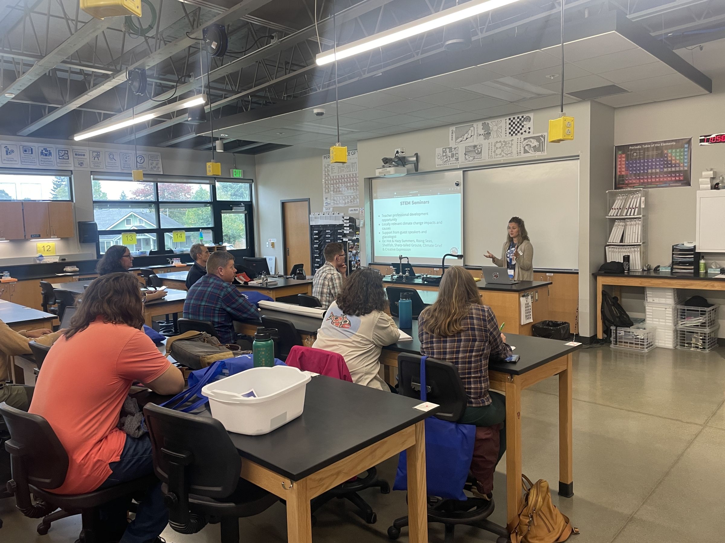 Bethany stands at the front of a classroom, presenting to several other teachers who are sitting in desks