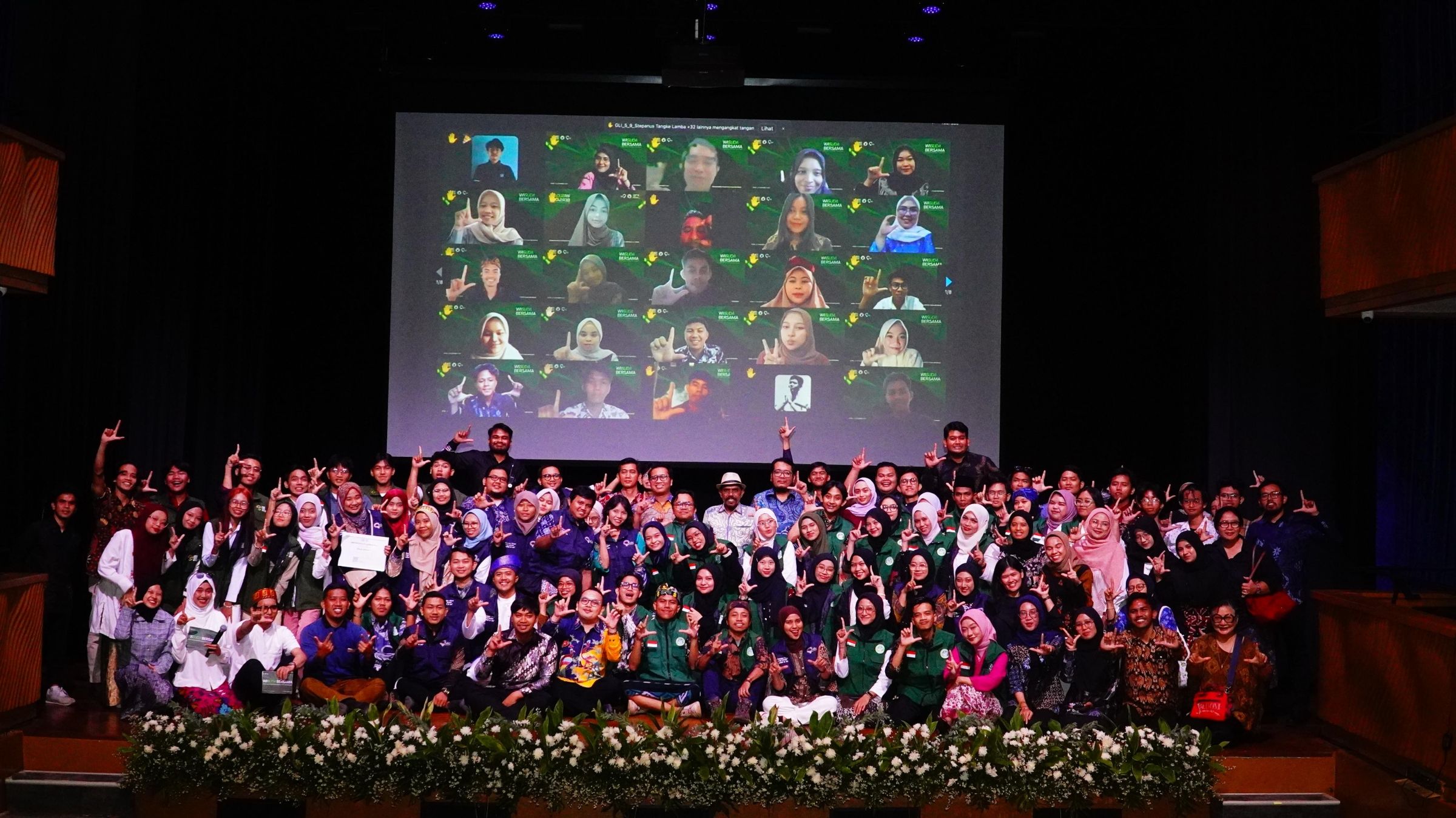 A large group of young people posing for a picture on a stage, with the faces of more young people joined virtually on a screen in the background