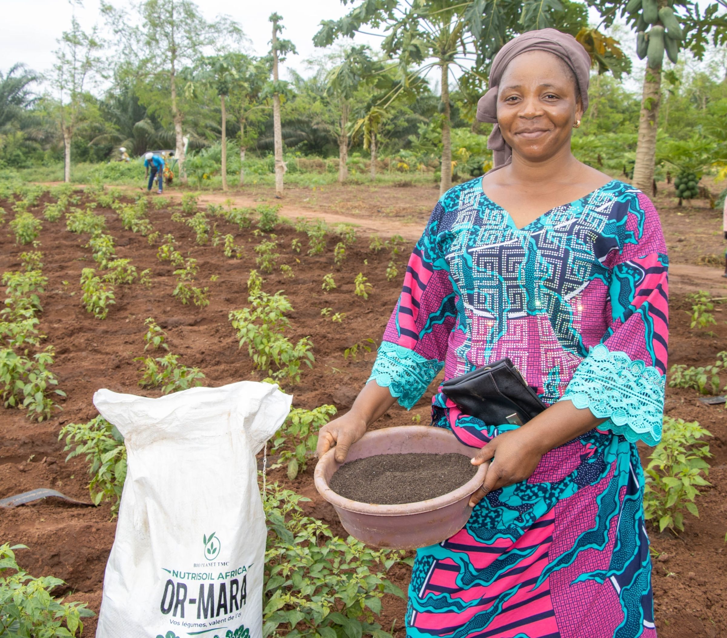 A woman poses for a picture while holding a pot of soil and standing in a newly planted field