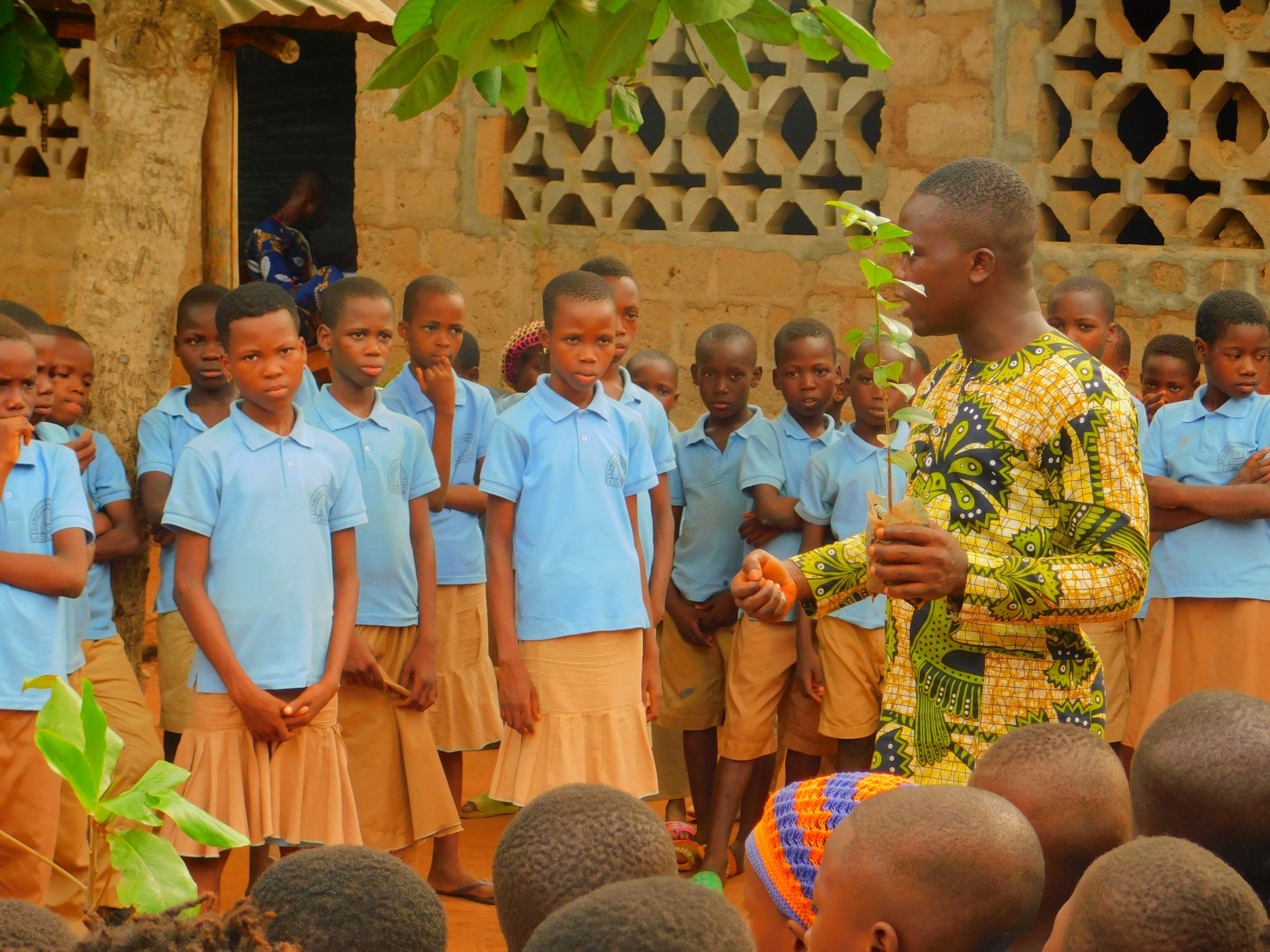 A young man holds a tree sapling and is presenting in front of a group of young students